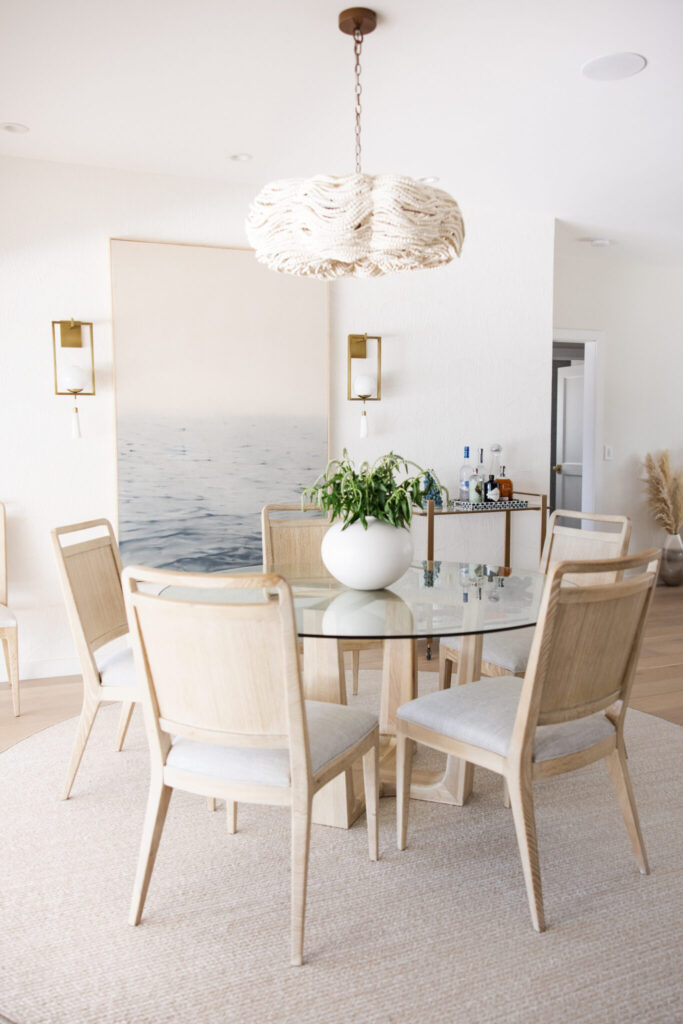 Modern coastal dining room with ocean-inspired artwork, glass table, and neutral textures designed by KH by Vesta Home.