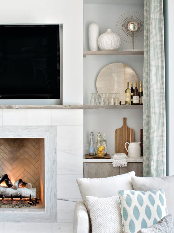 Cozy modern fireplace with marble surround, built-in shelving, bar accessories, and coastal textures in soft blue and white.