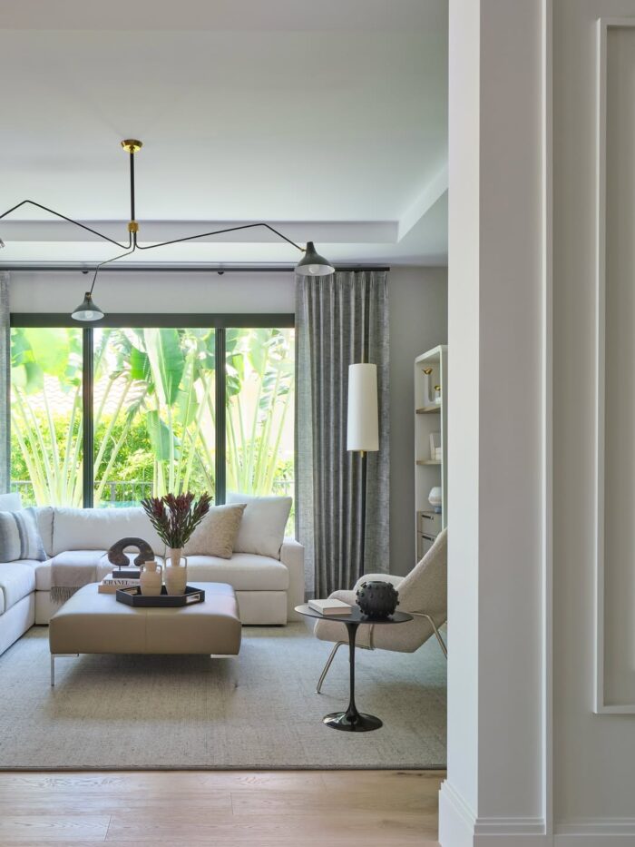 Modern living room with a white sectional sofa, taupe ottoman, gray drapery, large window overlooking tropical greenery, and soft neutral decor.