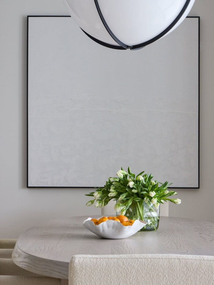 Minimalist dining table with a white ceramic bowl of oranges, a vase of white tulips, and a large monochromatic textured artwork behind it.