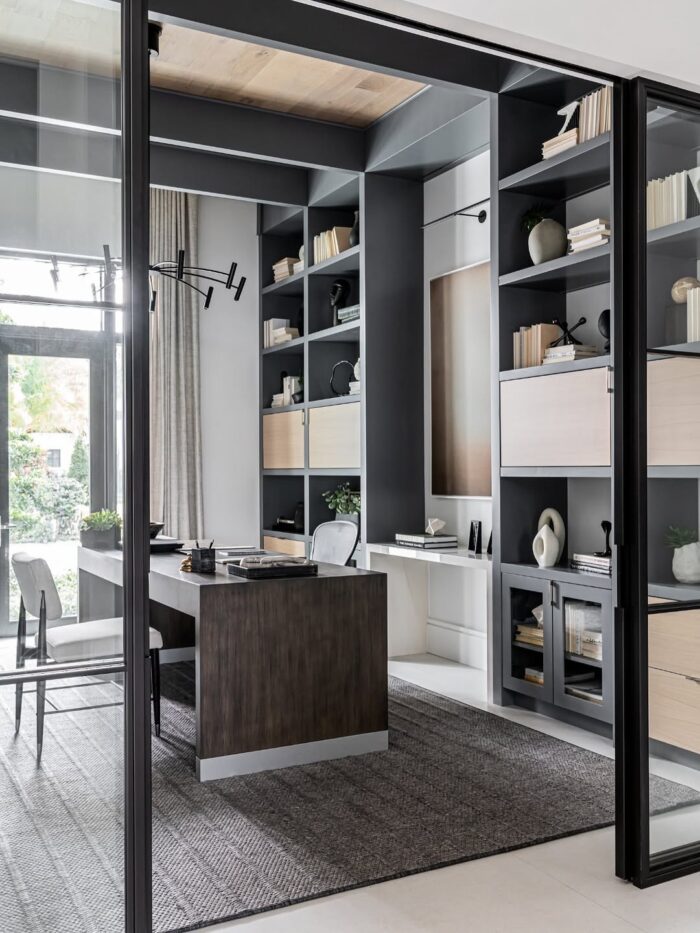 Modern home office with floor-to-ceiling built-in shelving, a large wood desk, glass wall partitions, and neutral furnishings. Soft natural light fills the room and a structured black light fixture hangs overhead.
