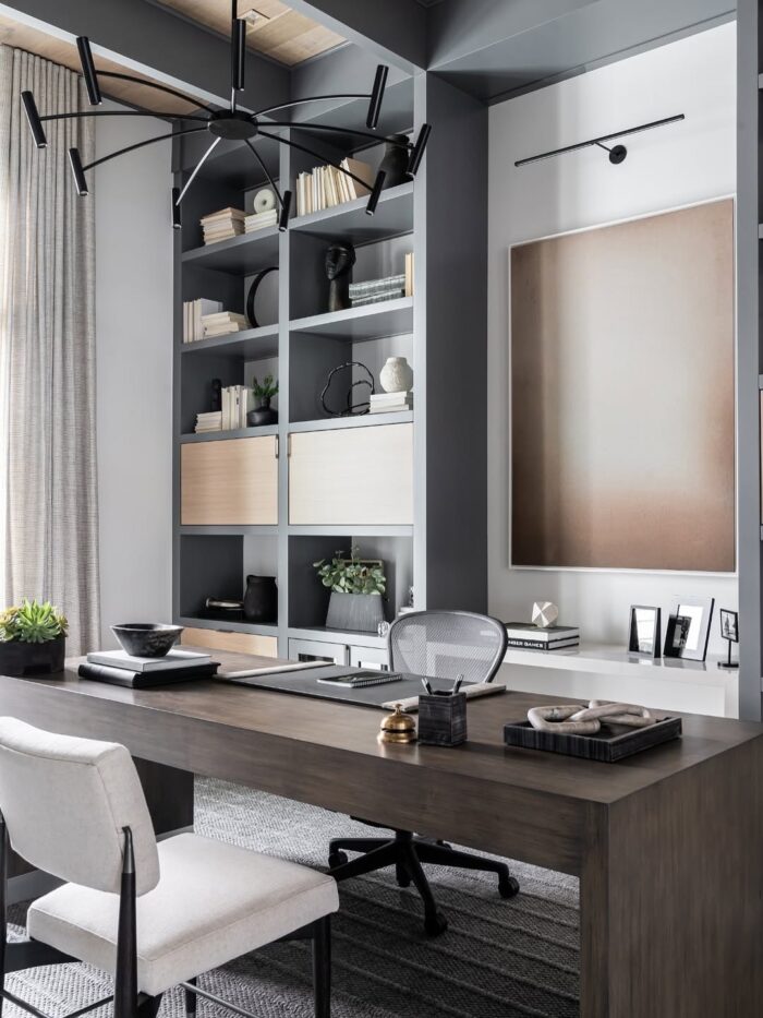 Contemporary office featuring custom gray built-ins, light wood cabinet fronts, abstract art, and a sleek modern desk. A mesh task chair and minimal accessories complete the clean workspace.