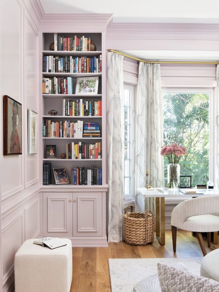 Cozy reading nook with blush-painted built-in bookshelves, brass hardware, a white desk with gold legs, floral curtains, and a woven basket beneath the window.