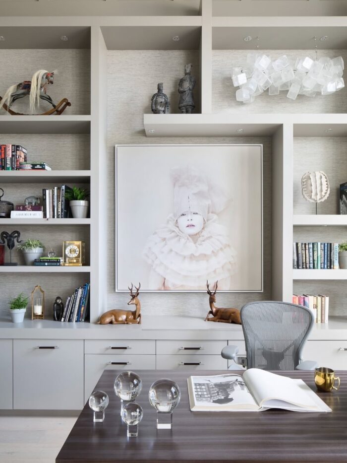 Sophisticated office with light gray built-ins, a large framed art piece, wood desk, decorative sculptures, and glass spheres styled on the desktop.