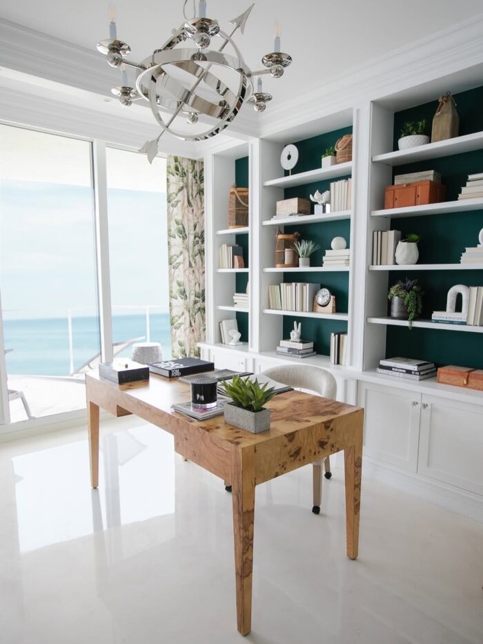 Bright coastal home office with ocean views, white built-ins, deep teal back panels, a natural wood desk, and modern chrome chandelier. Tropical curtains flank the large window.